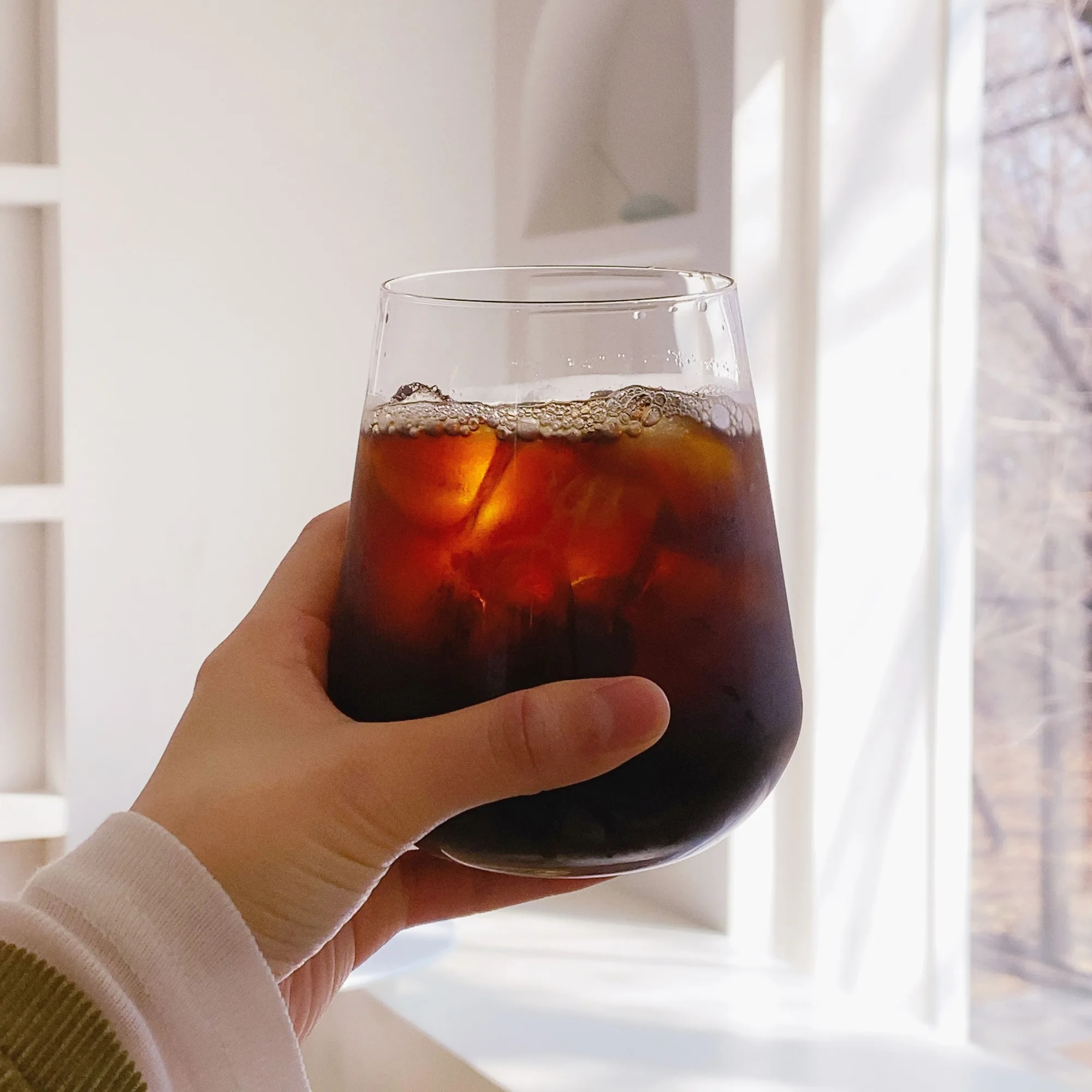 Hand holding a glass of iced Americano coffee with visible ice cubes in bright natural light