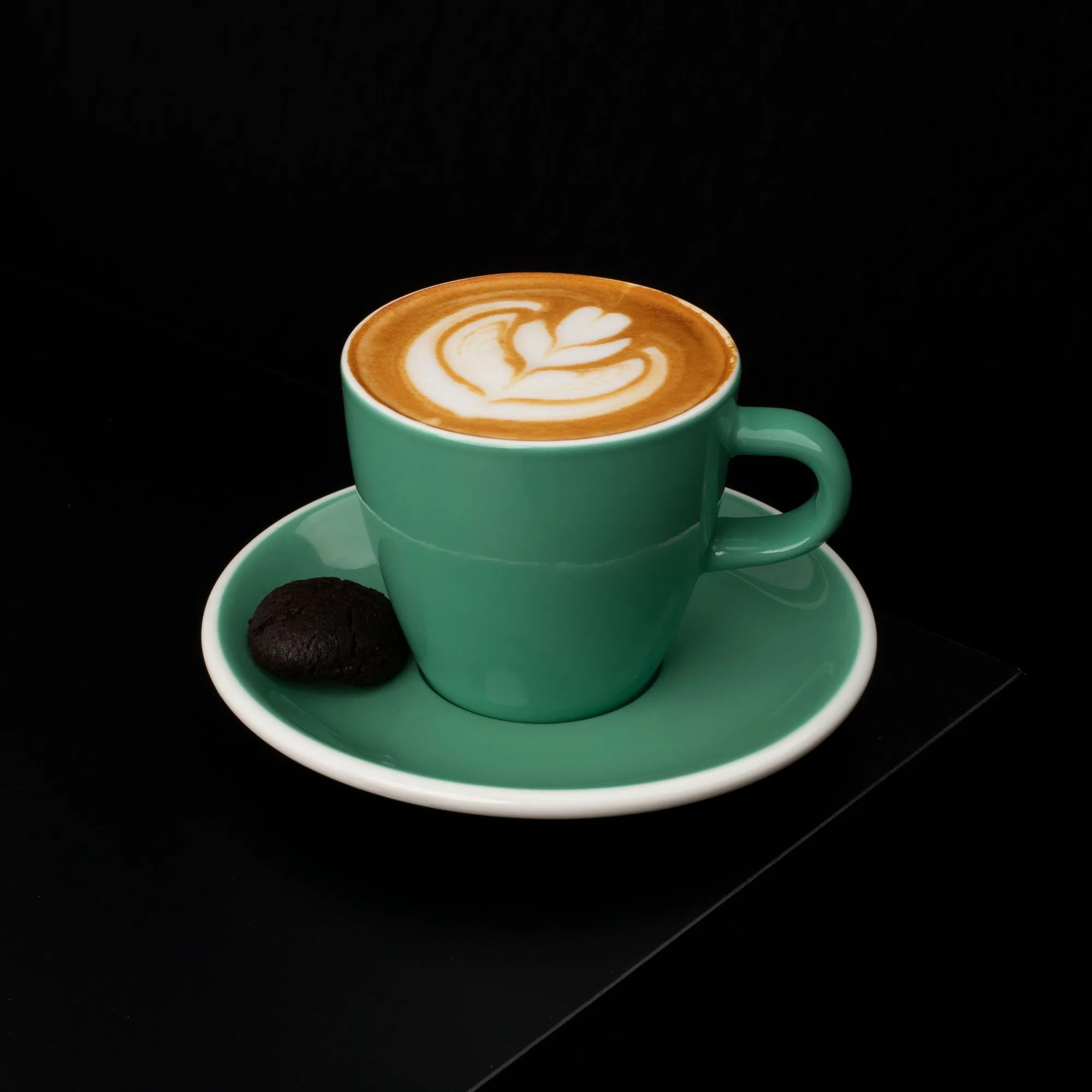 A cortado with latte art in a small green ceramic cup beside a biscuit on a dark surface