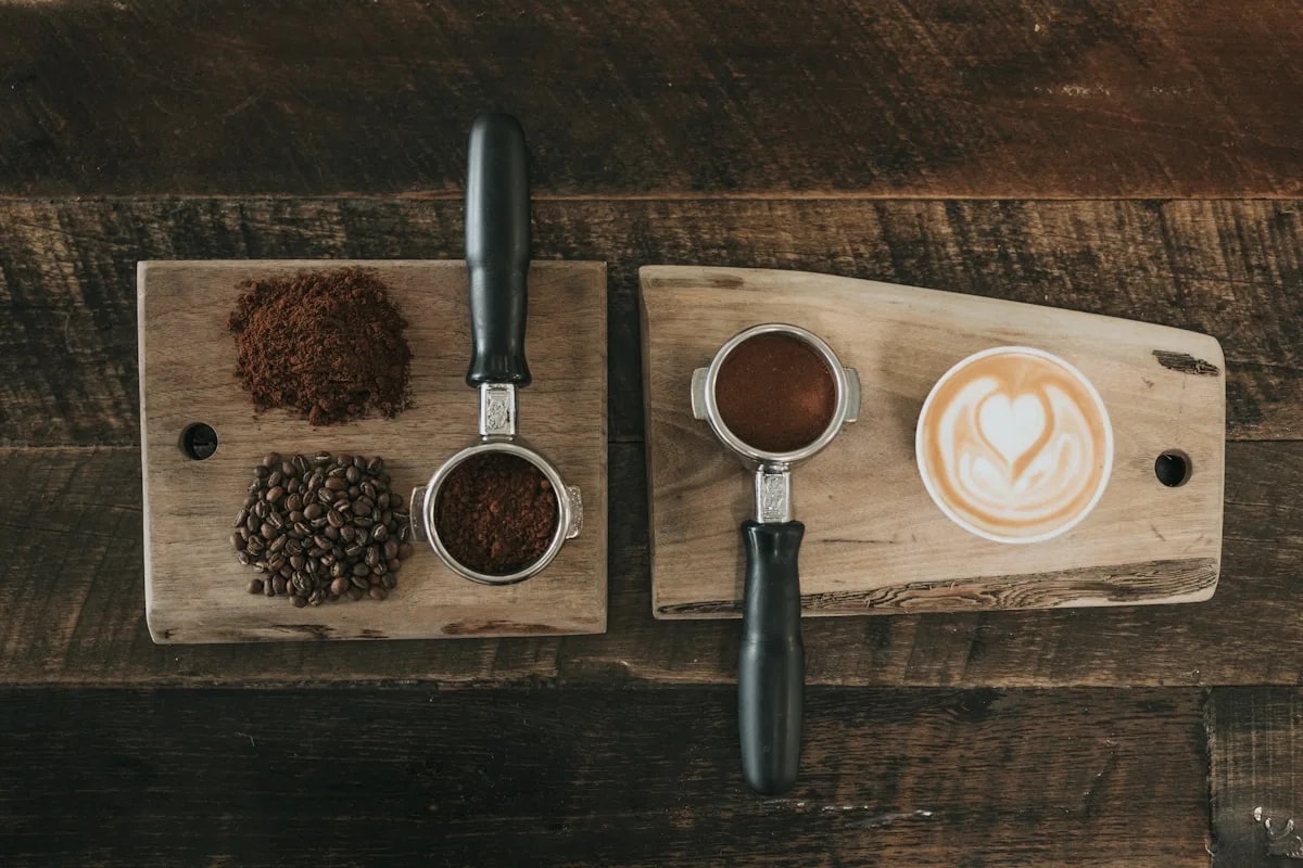 Overhead view of a wooden board with a portafilter, coffee grounds, whole beans, and a cup of latte with heart-shaped latte art