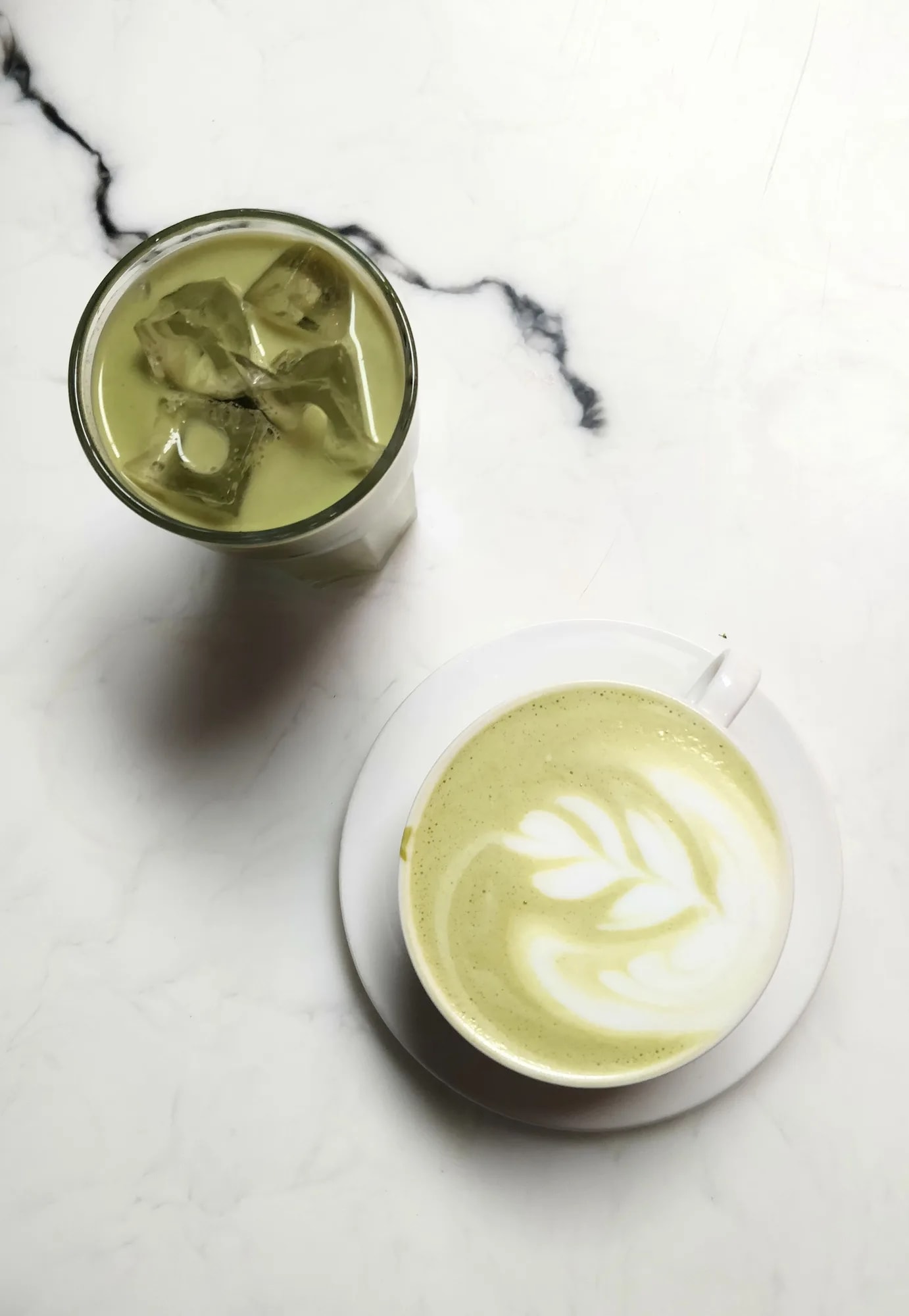 Overhead view of a matcha latte with latte art beside a glass of iced green tea on a marble table