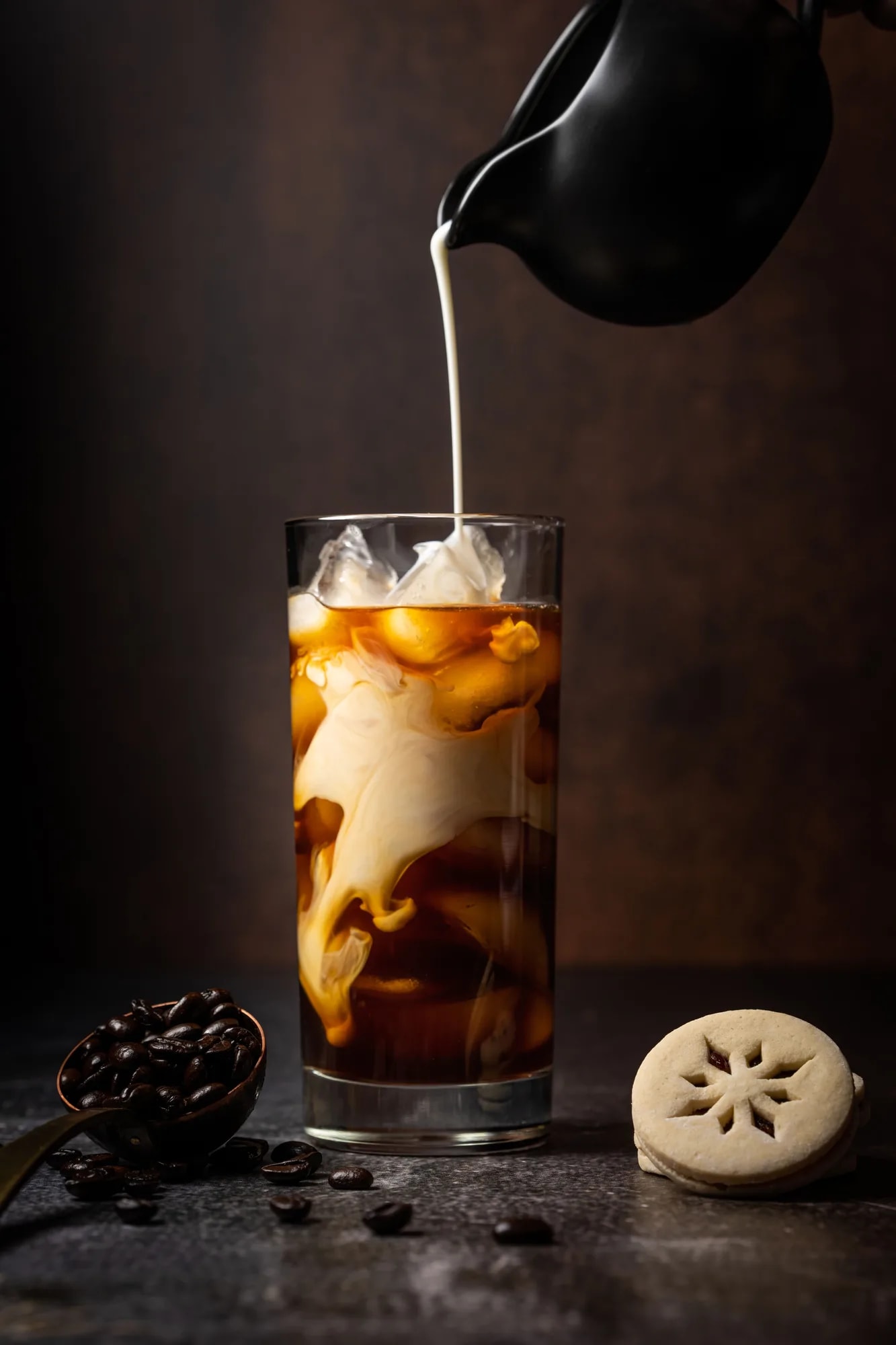 Milk being poured into iced coffee in a tall glass, creating a two-tone layered effect, with coffee beans nearby
